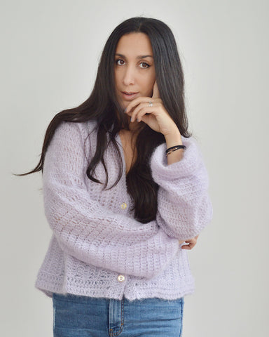Woman with dark hair and arms crossed, wearing lilac mohair crochet cardigan and blue jeans