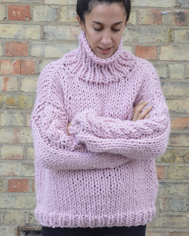 Woman with arms crossed wearing pink chunky knit jumper against brick background