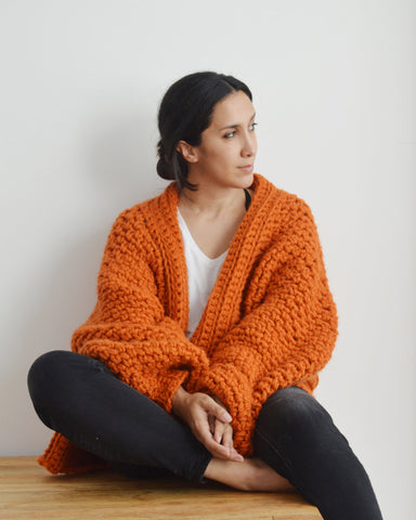 Woman seated wearing orange chunky crochet cardigan, black jeans and white tee against white background