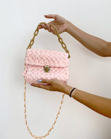 Pale pink knitted handbag with gold chain held by woman’s arms against white wall