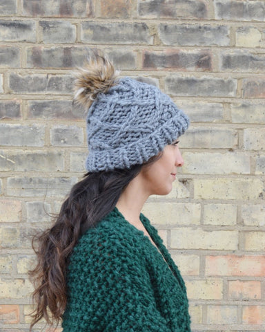 Woman standing side on wearing grey knit beanie with brown pompom