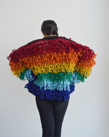 Back view of woman standing with hands on hips wearing knitted loopy cardigan in rainbow shades