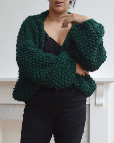 Woman wearing green chunky knit cardigan and black jeans, with one arm crossed at waist