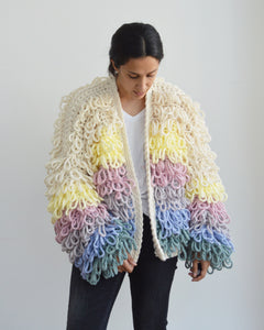 Woman looking down wearing loopy pastel crochet cardigan against light background